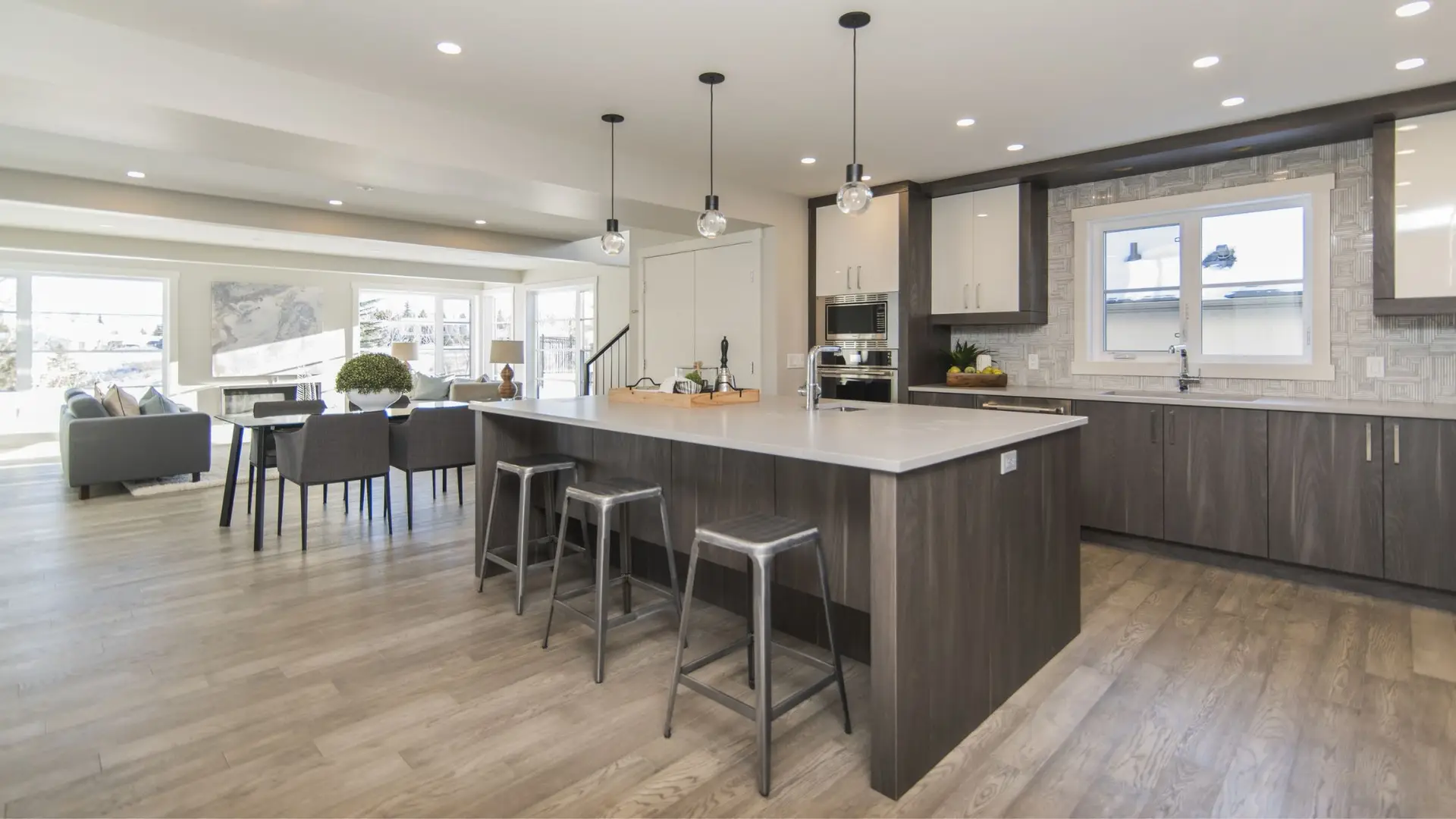 7-Surprising-Benefits-of-Kitchen-Remodeling Modern kitchen with gray cabinets, a large island, stainless steel stools, and a bright open layout featuring natural light and contemporary decor.