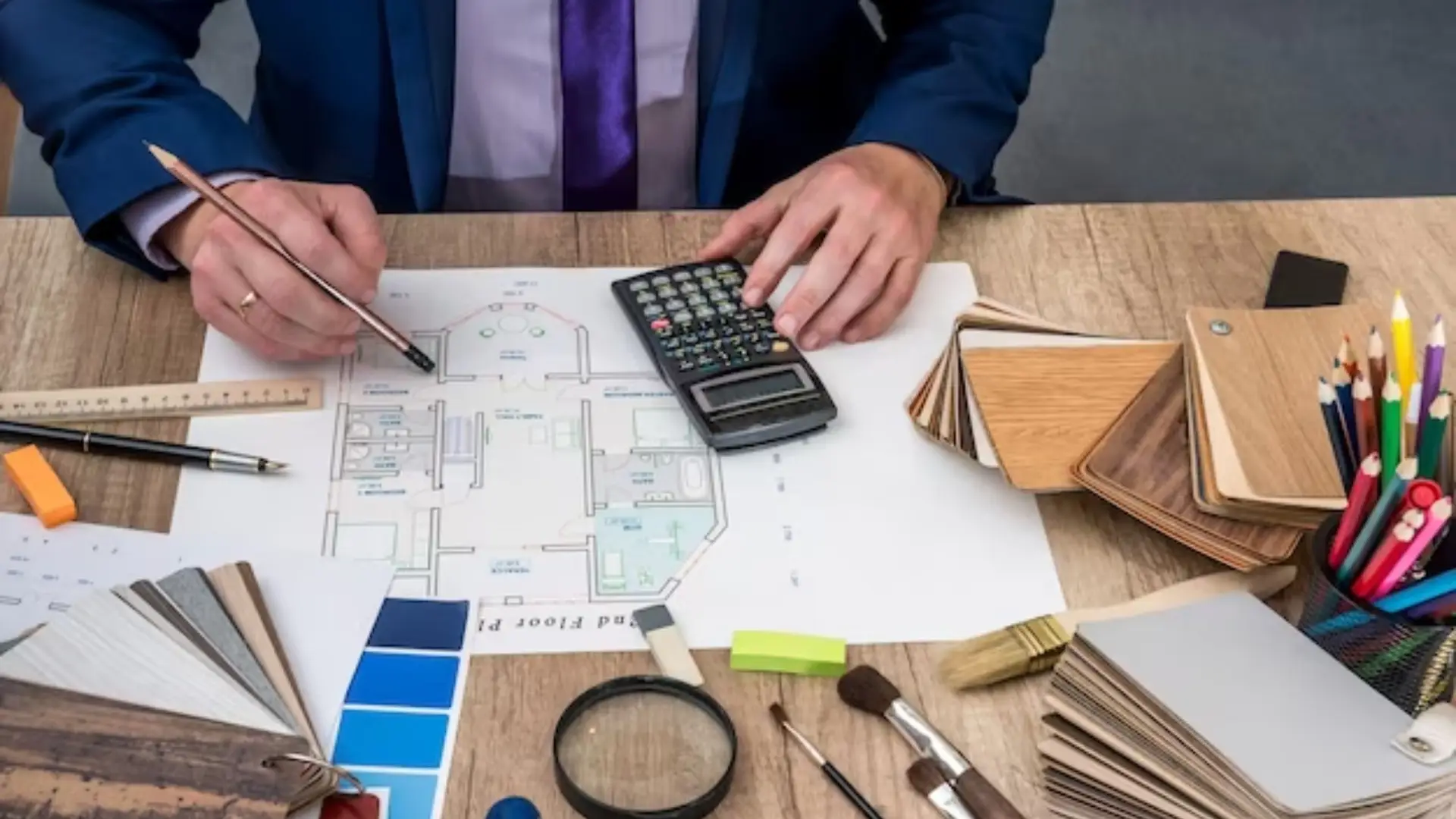 Person analyzing kitchen remodeling plans with a calculator, design sketches, color swatches, and tools on a wooden desk.